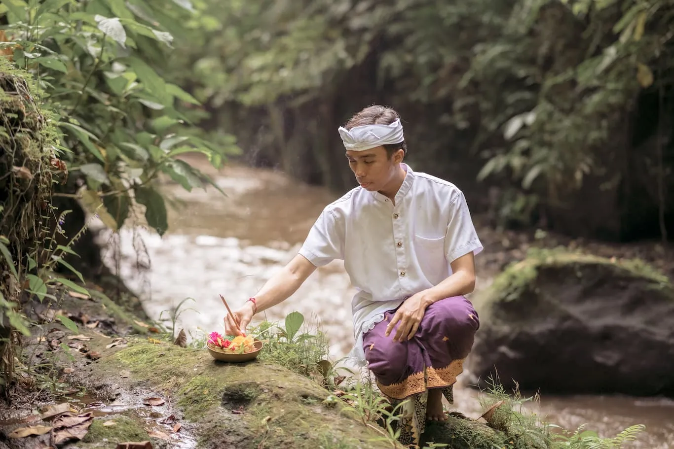 A man in ceremonial dress places an offering of flowers on a rock beside the river.