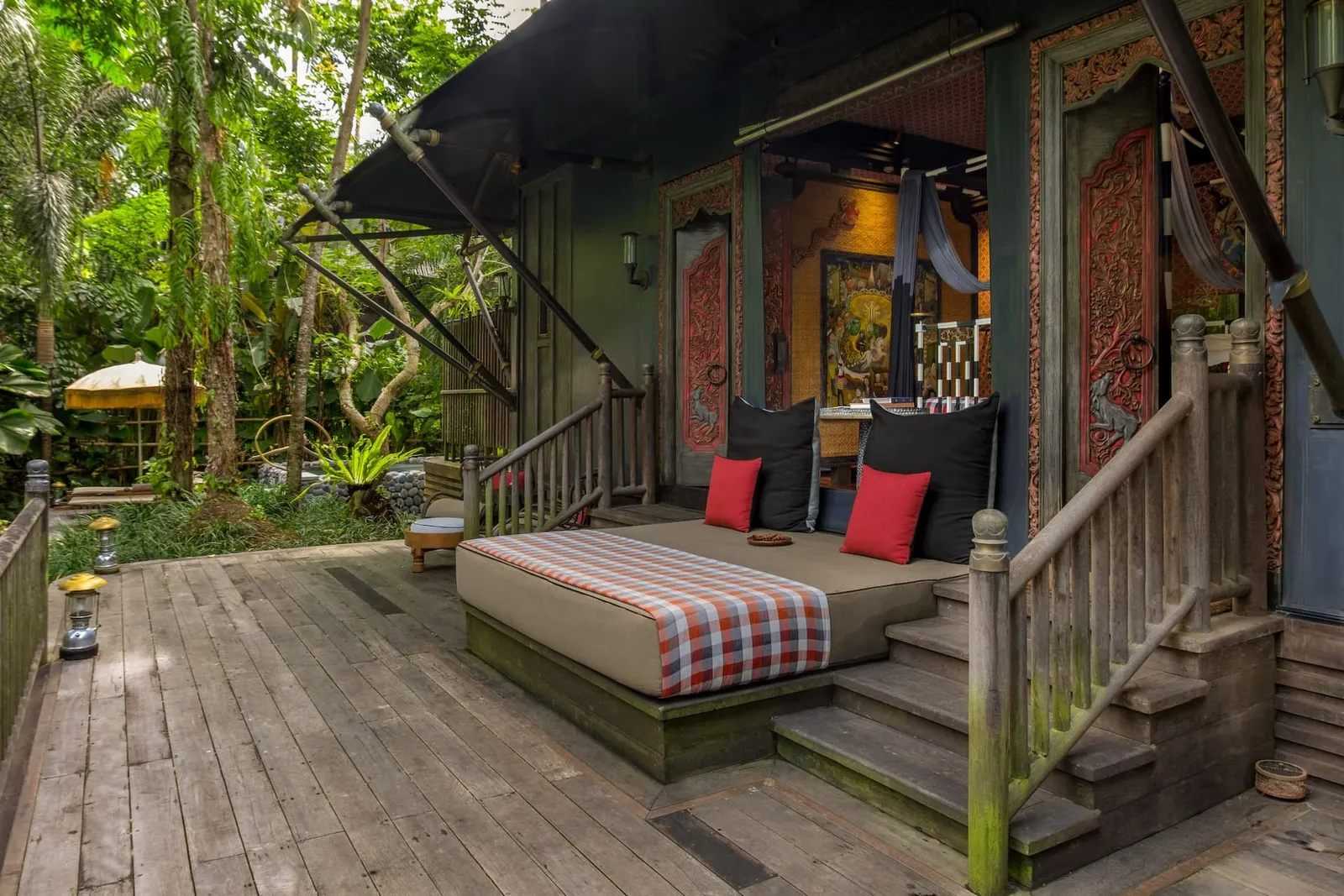 A Forest Tent’s deck: the daybed with red cushions, the wooden steps up to the tent and the garden among the trees.