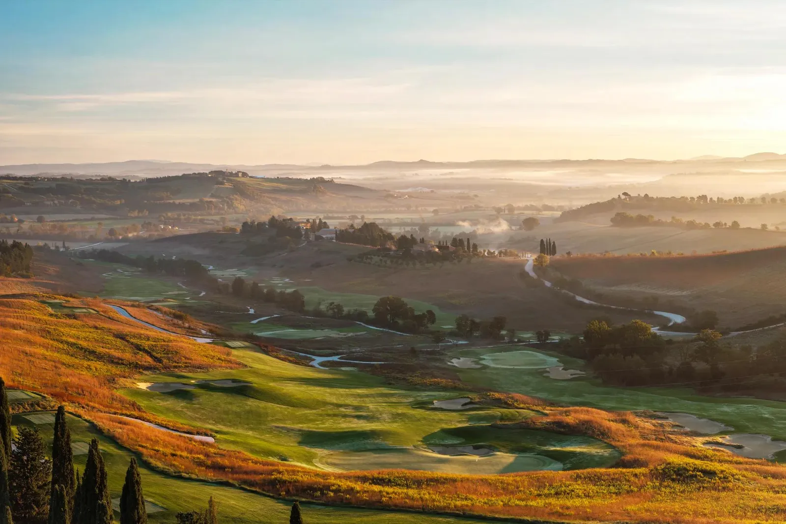 The golf course at dawn, with mist in the valley, bunkers and cypresses and the hills in the distance.