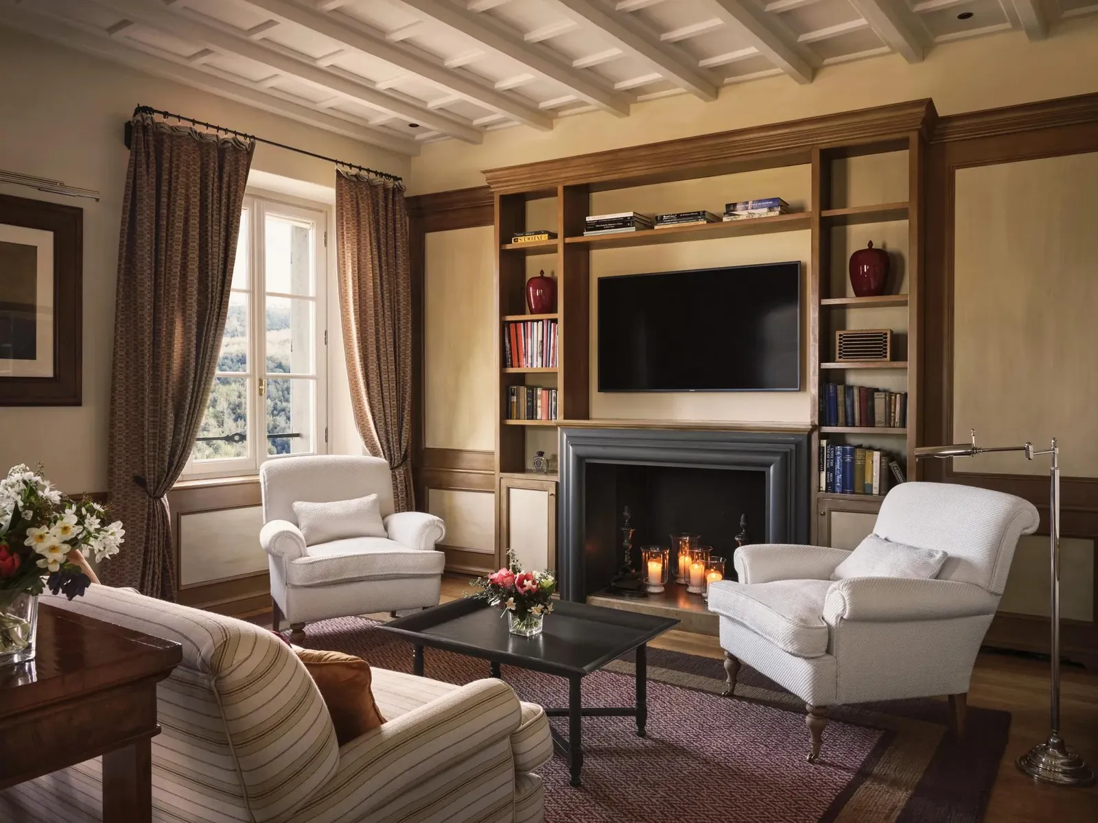 A Borgo Suite’s living room, with the fire lit, the bookcase, 2 white armchairs and a striped sofa.