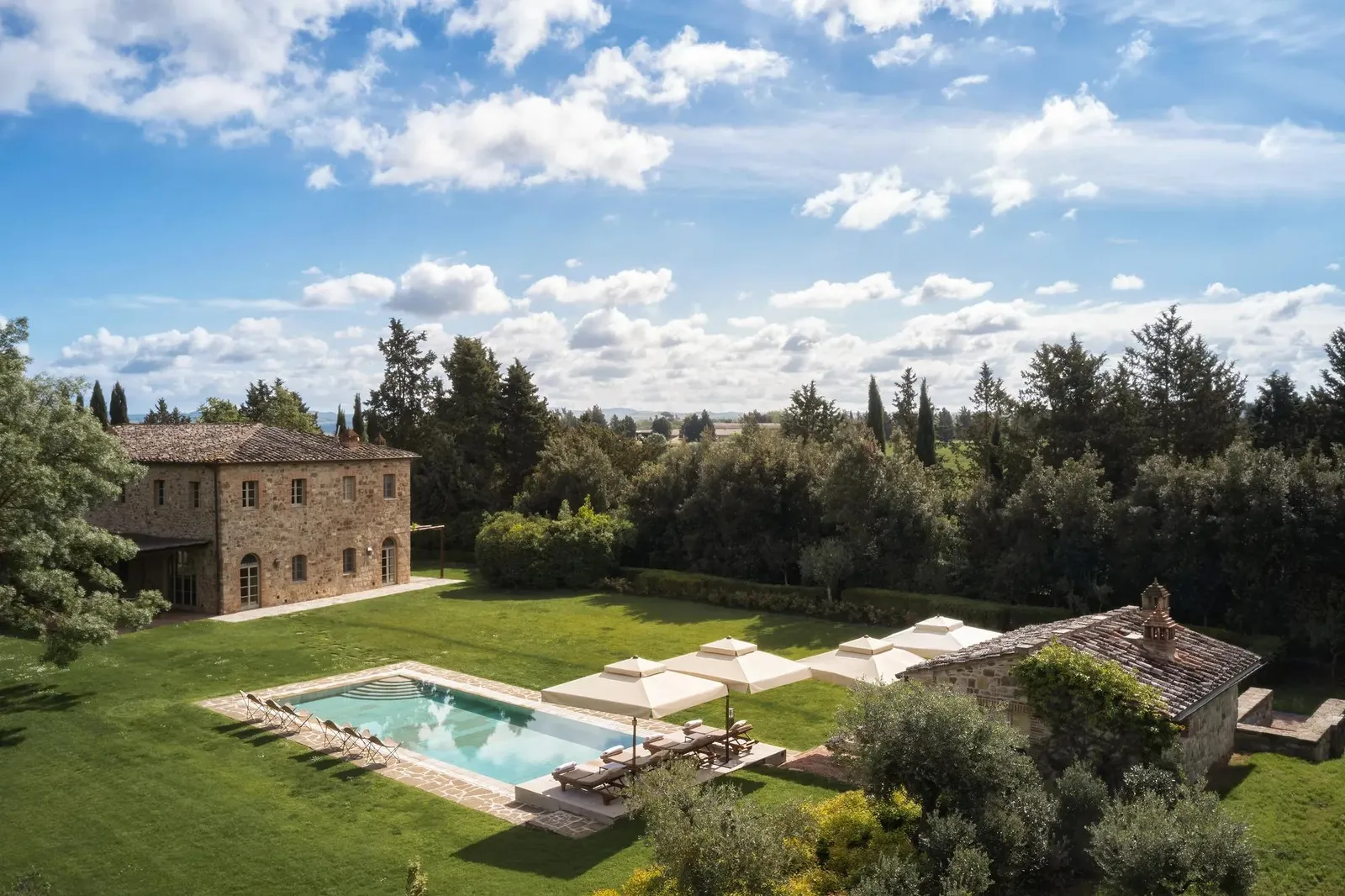 Villa Biondi among lawns and trees, with its pool, white loungers and the stone house behind.