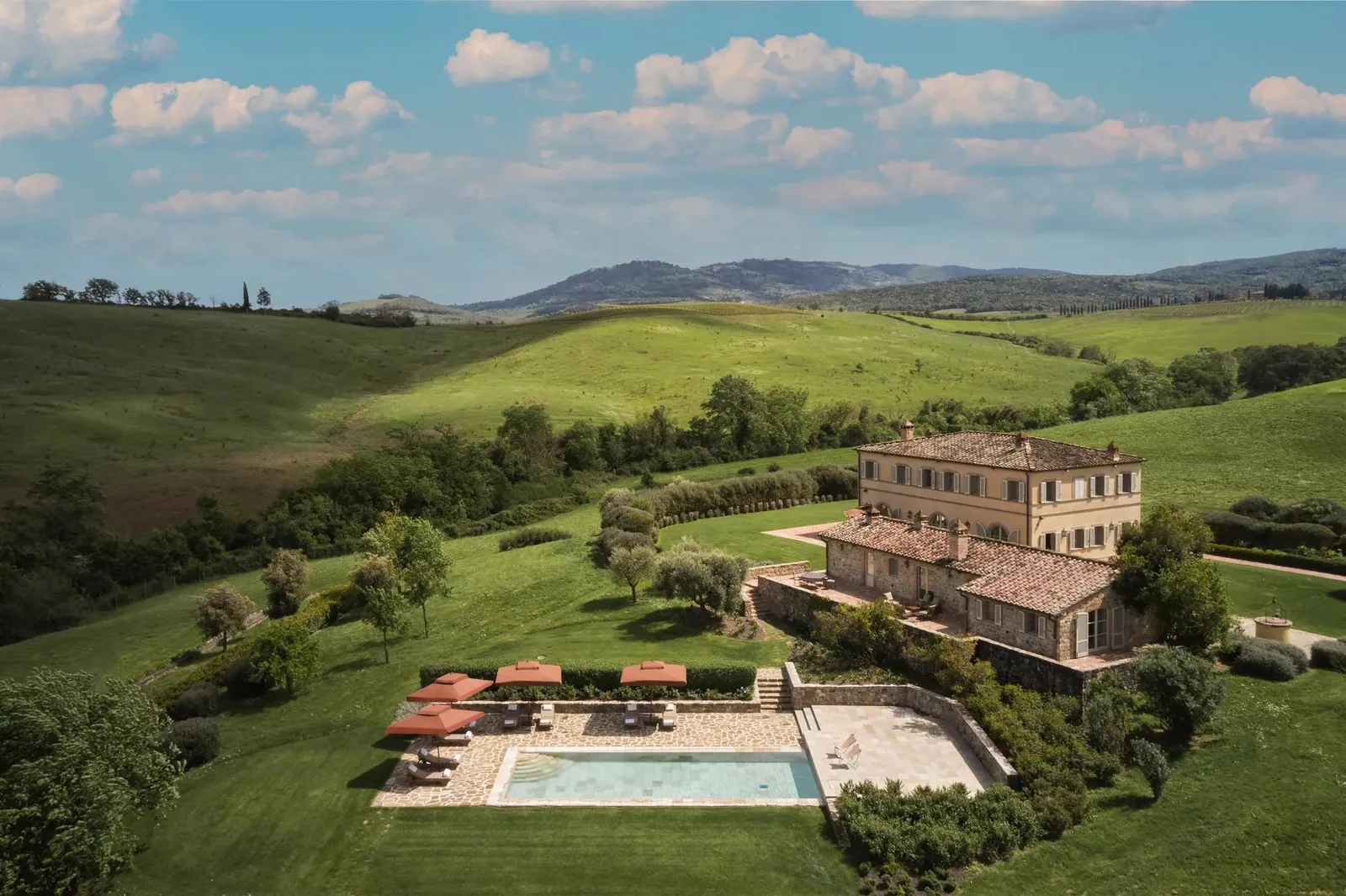 Villa Casa del Fiume from above, in a valley of meadows: the stone house, the pool and the red parasols.