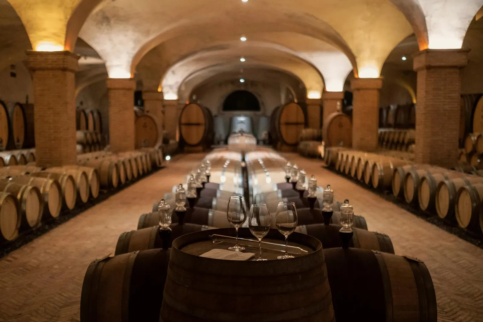The winery’s barrel room under brick vaults: 2 rows of oak casks and a table laid with glasses down the middle.