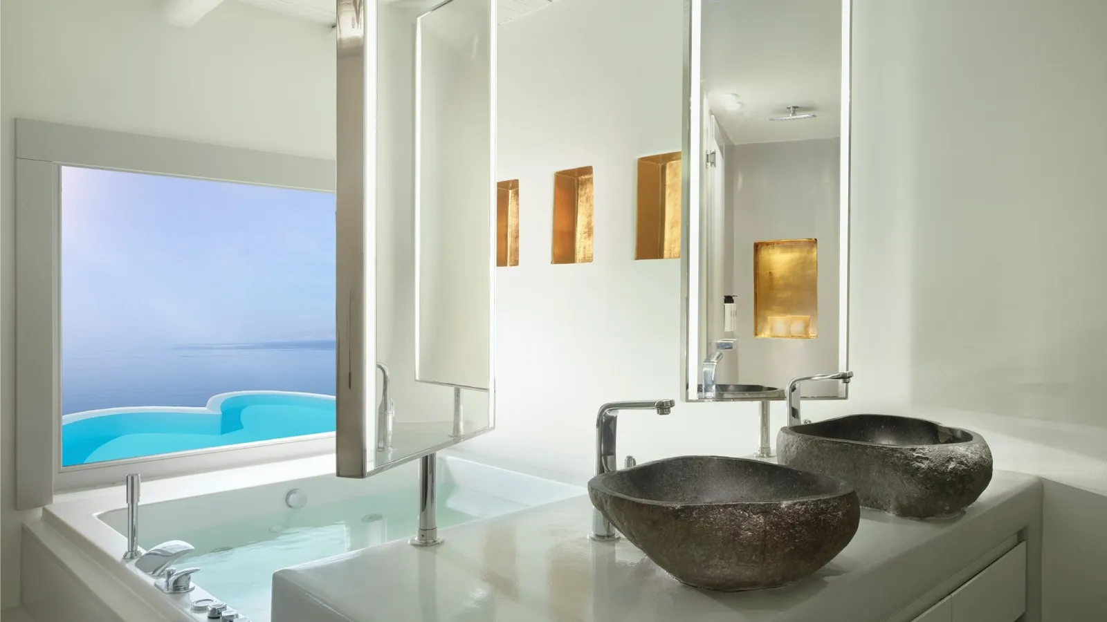 A bathroom with 2 rough stone basins, a bath and a window onto a heart-shaped pool and the sea.