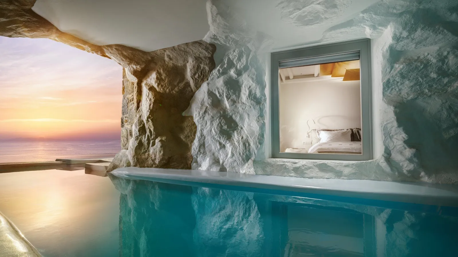 A pool cut into whitewashed rock, a bedroom window looking onto the water and the sea at sunset beyond the opening.