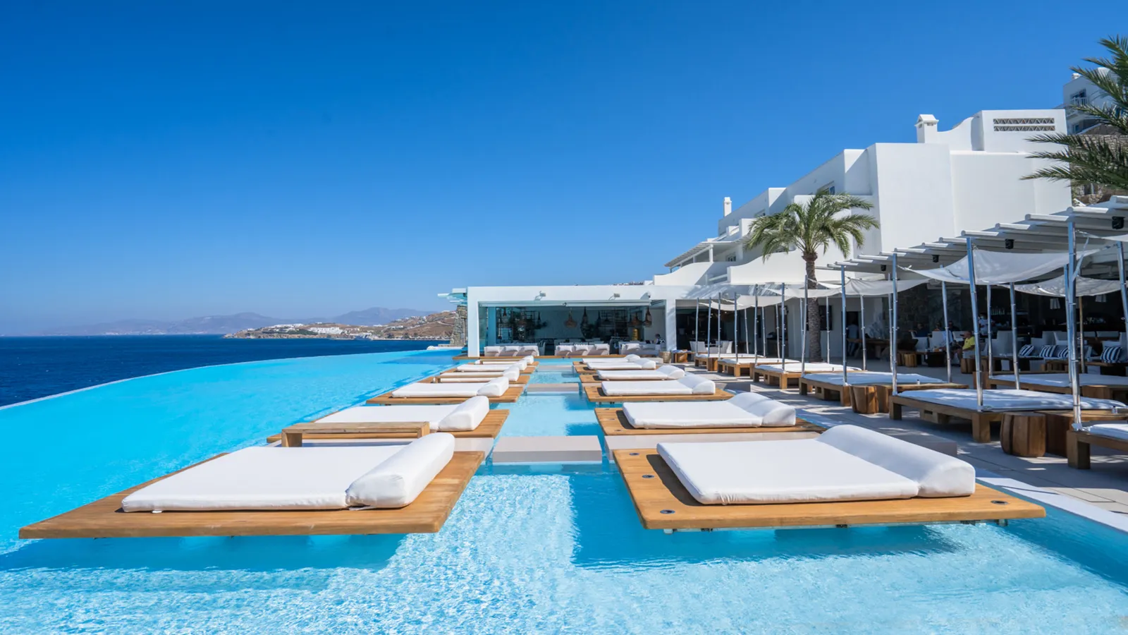 The infinity pool by day, with white daybeds on timber decks set in the water, the lounge pavilion and the hotel’s white buildings.