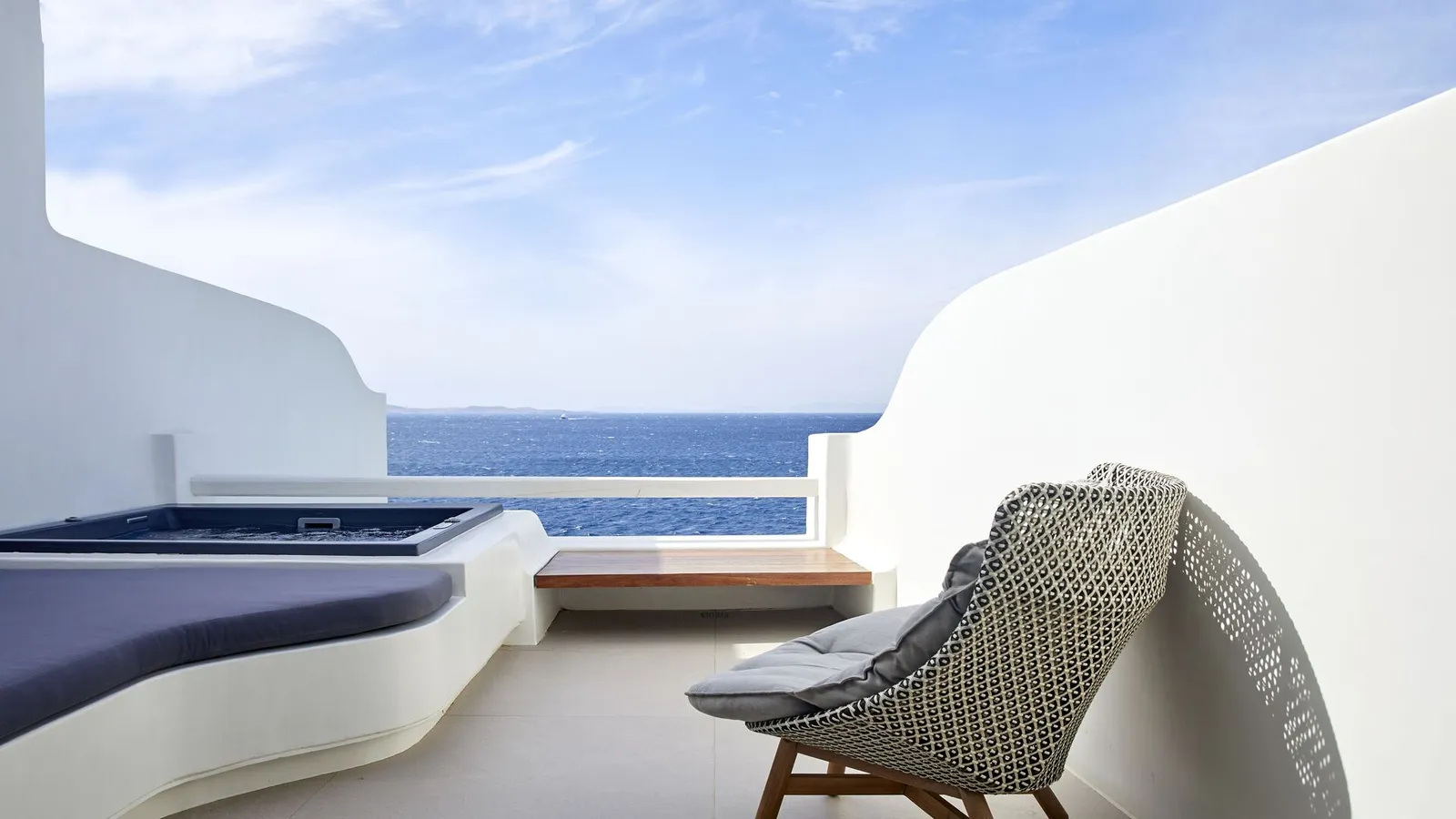 A terrace between curved white walls, with a hot tub, a wicker chair and the sea above the parapet.