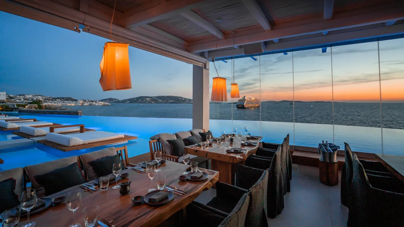 A laid timber table at Meraki under orange lanterns, with the pool’s daybeds and Mykonos bay at dusk.