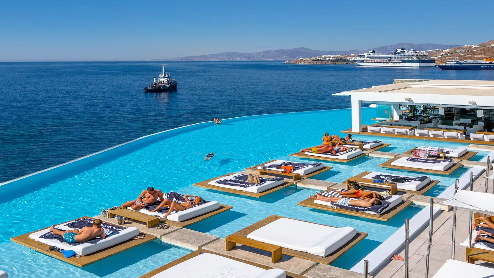 The pool from above on a summer day, guests on the daybeds, the bar and, in the bay, a tug and a cruise ship.