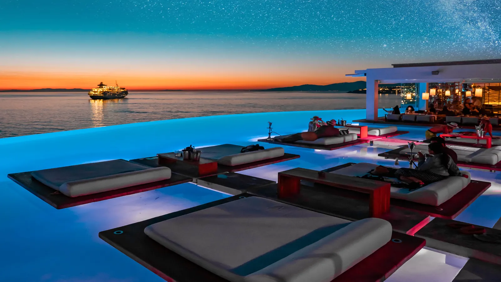 The pool after sunset: daybeds on lit decks, the restaurant glowing on the right and a ferry crossing the bay.