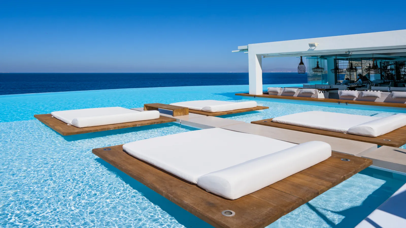 White double daybeds on timber decks in the middle of the pool, its infinity edge on the Aegean and the bar pavilion behind.