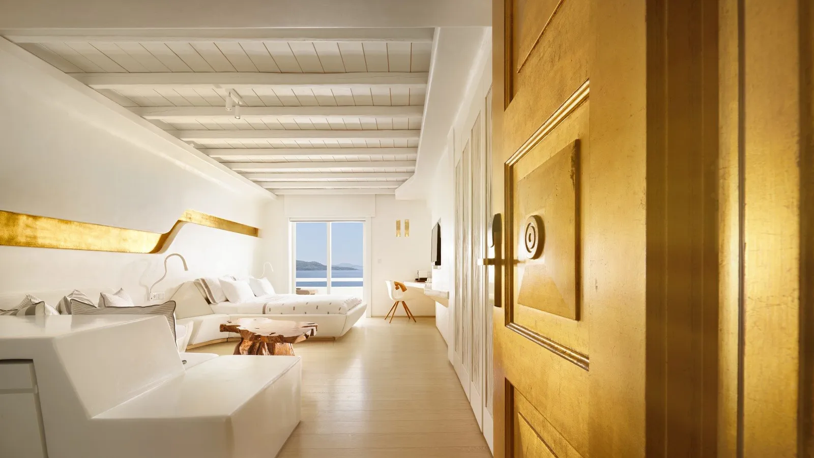 A Premium Room seen from its gilded door: the bed at the far end, a white sofa, a beamed ceiling and the window on the sea.