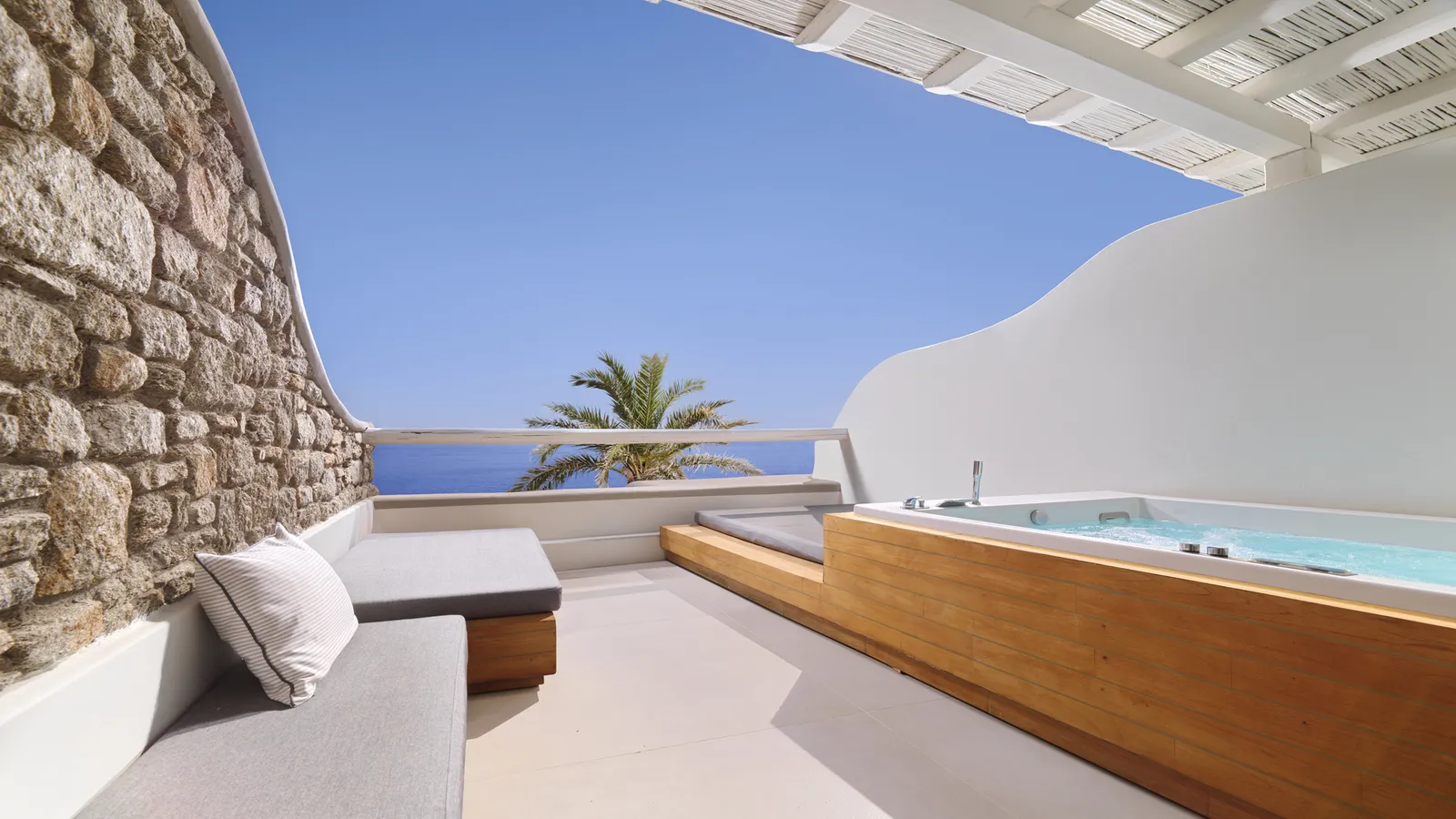 The terrace of the 2-bedroom suite: a timber-clad hot tub, a cushioned bench, a stone wall and a palm.