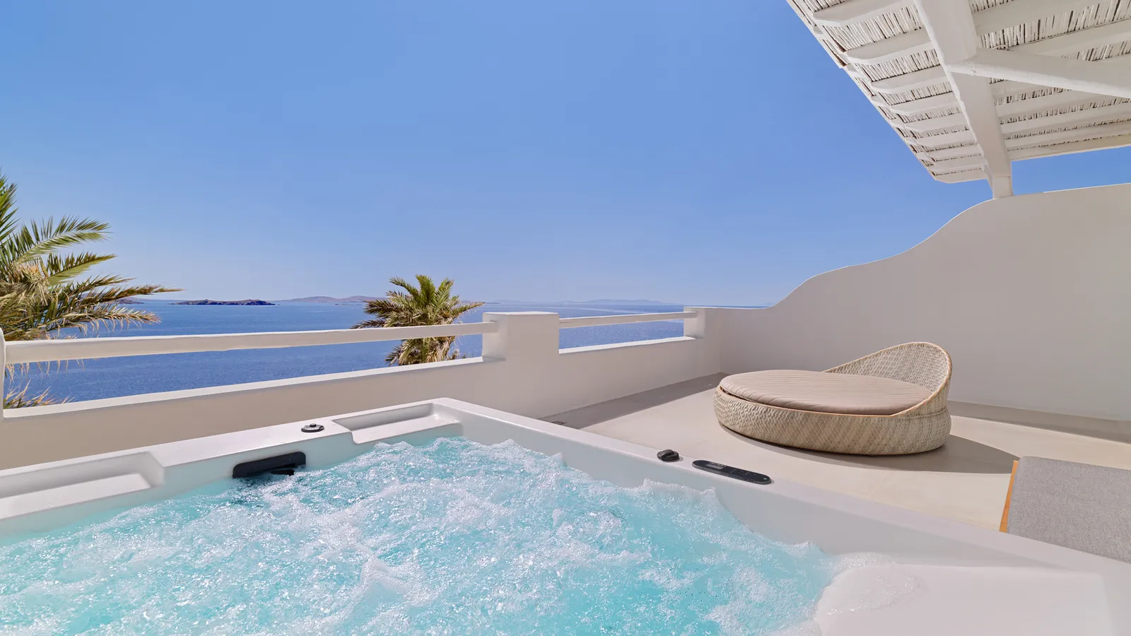 The terrace of a Suite with Hot Tub: the hot tub in the foreground, a wicker daybed, palms and the sea.