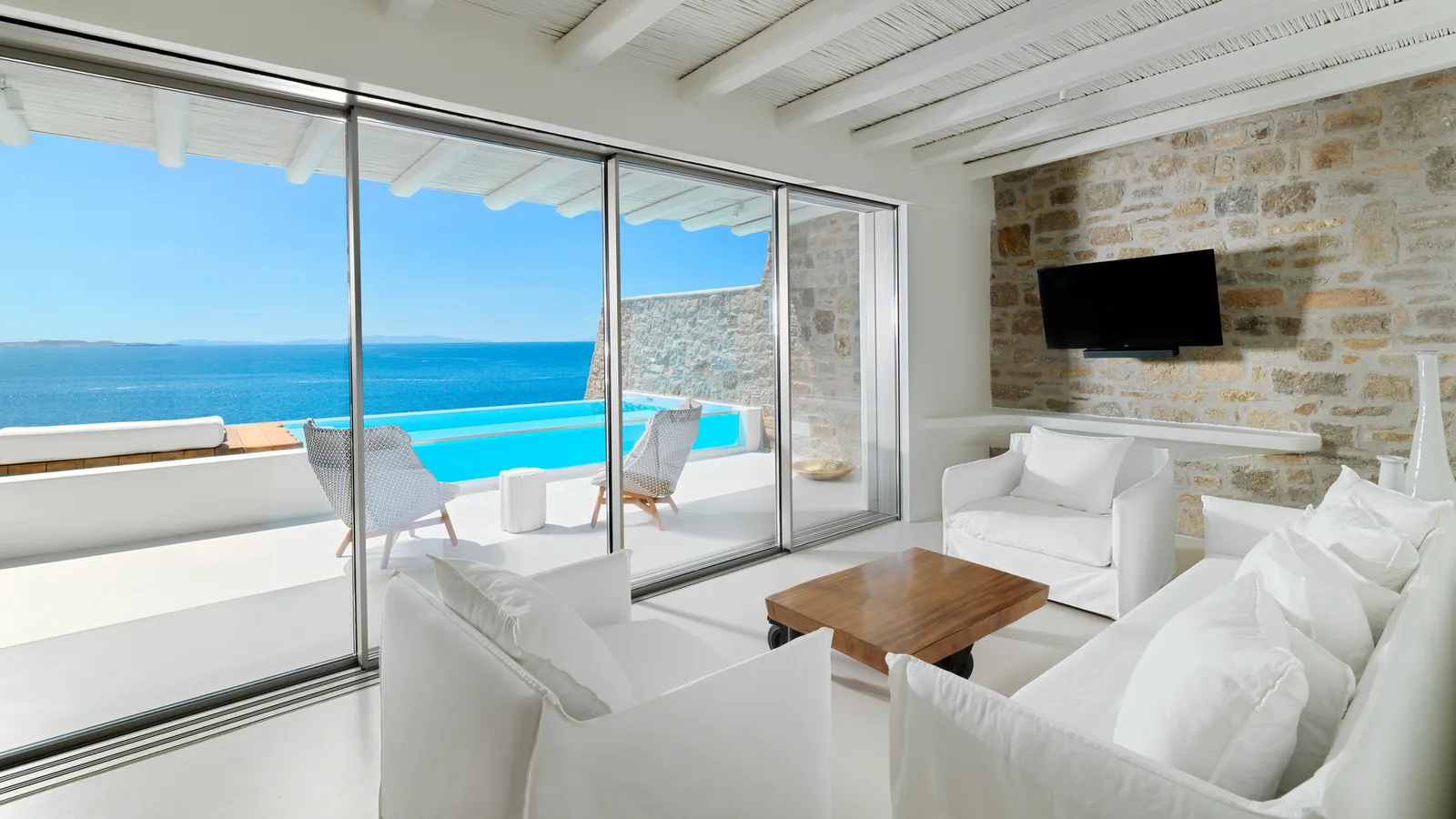 The living room of a Suite with Private Pool, with white sofas, a stone wall and glass doors open to the pool and the sea.