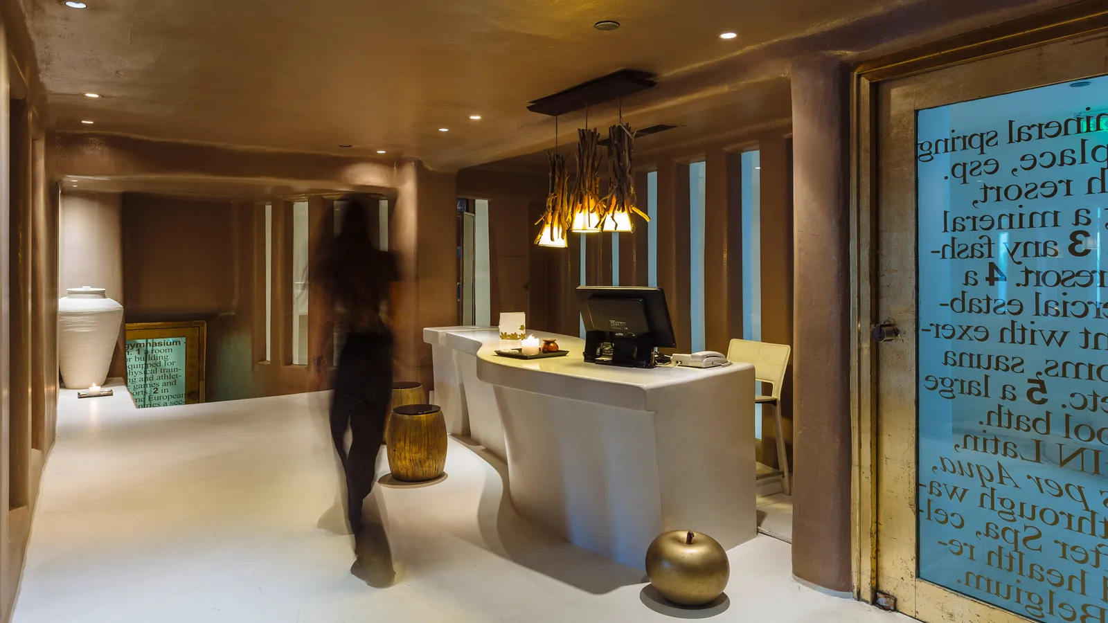 The spa’s entrance, with a masonry reception desk, low lights, an etched glass door and a figure walking through.