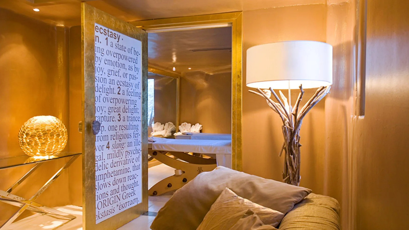 The spa: gilded walls, a glass door covered in lettering, a lamp on a base of twisted branches and a massage bed beyond.