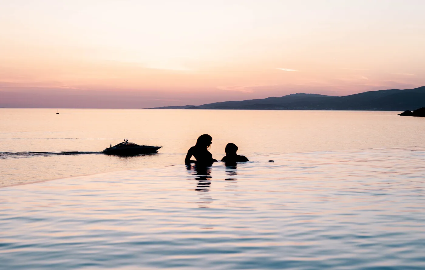 2 people in silhouette in the infinity pool at sunset, a speedboat passing and the coast beyond.