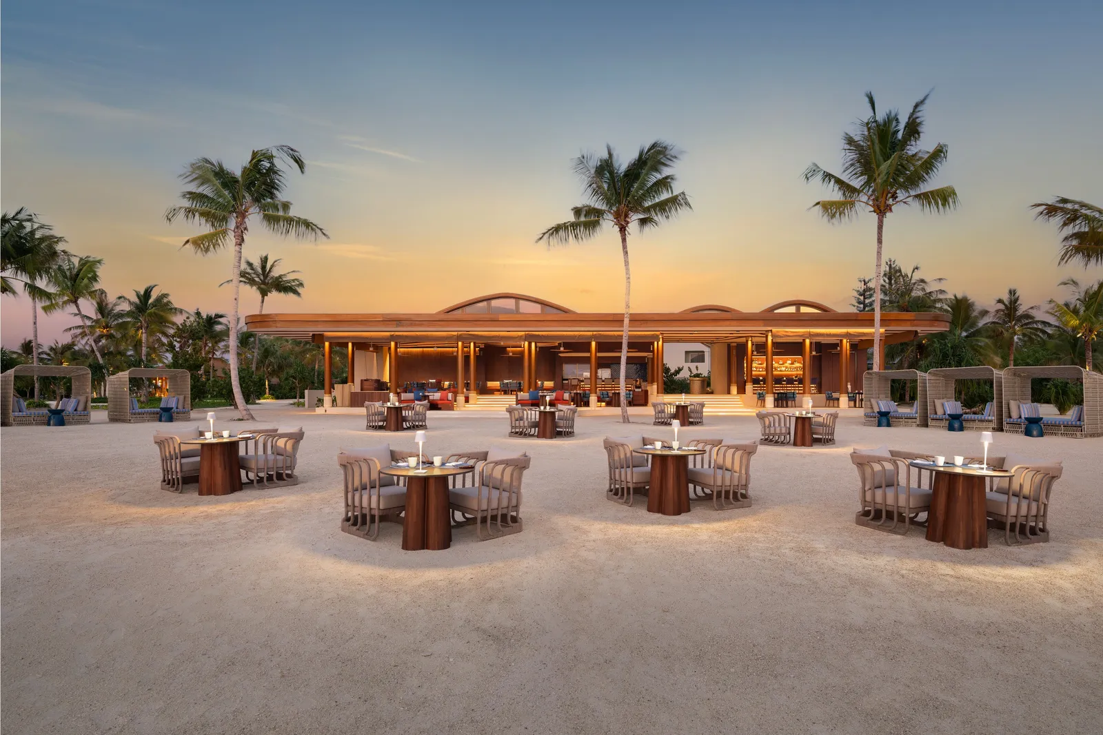 Bluefin’s tables on the sand at dusk, among the palms, the pavilion lit behind