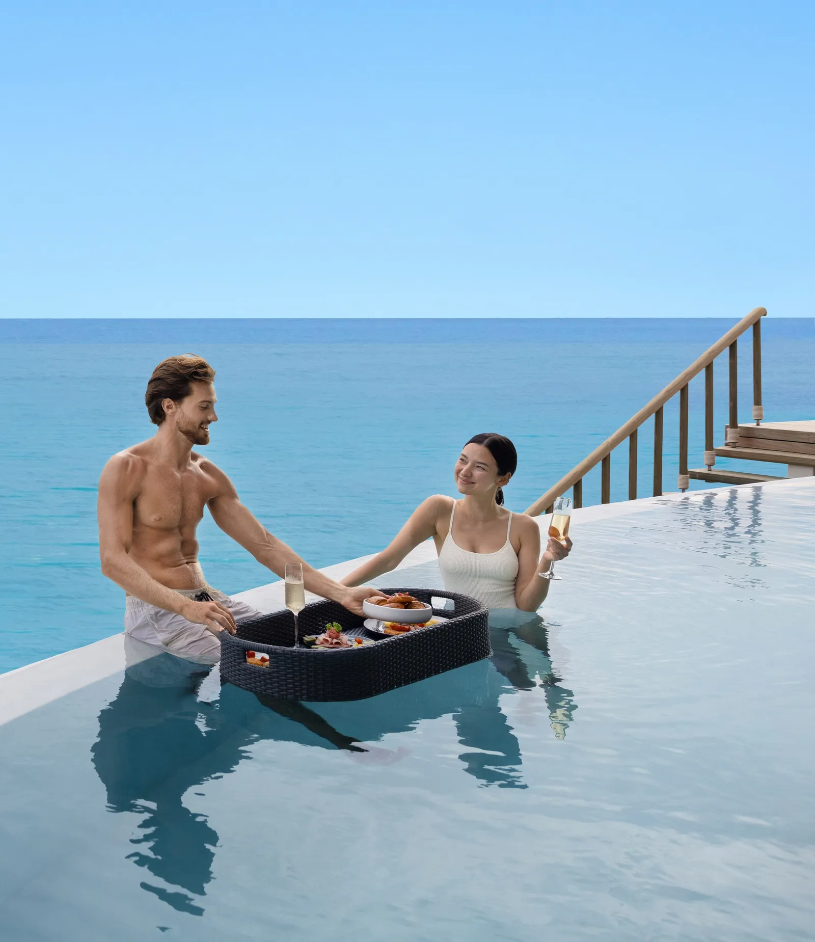 A couple in their villa pool with the breakfast tray floating beside them, the sea behind