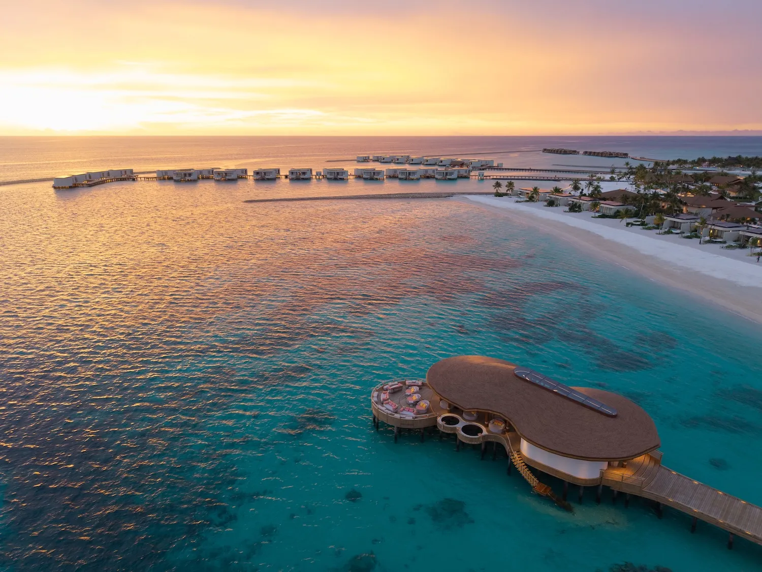Sunset over the lagoon, an overwater pavilion with cushions on its deck and the row of villas