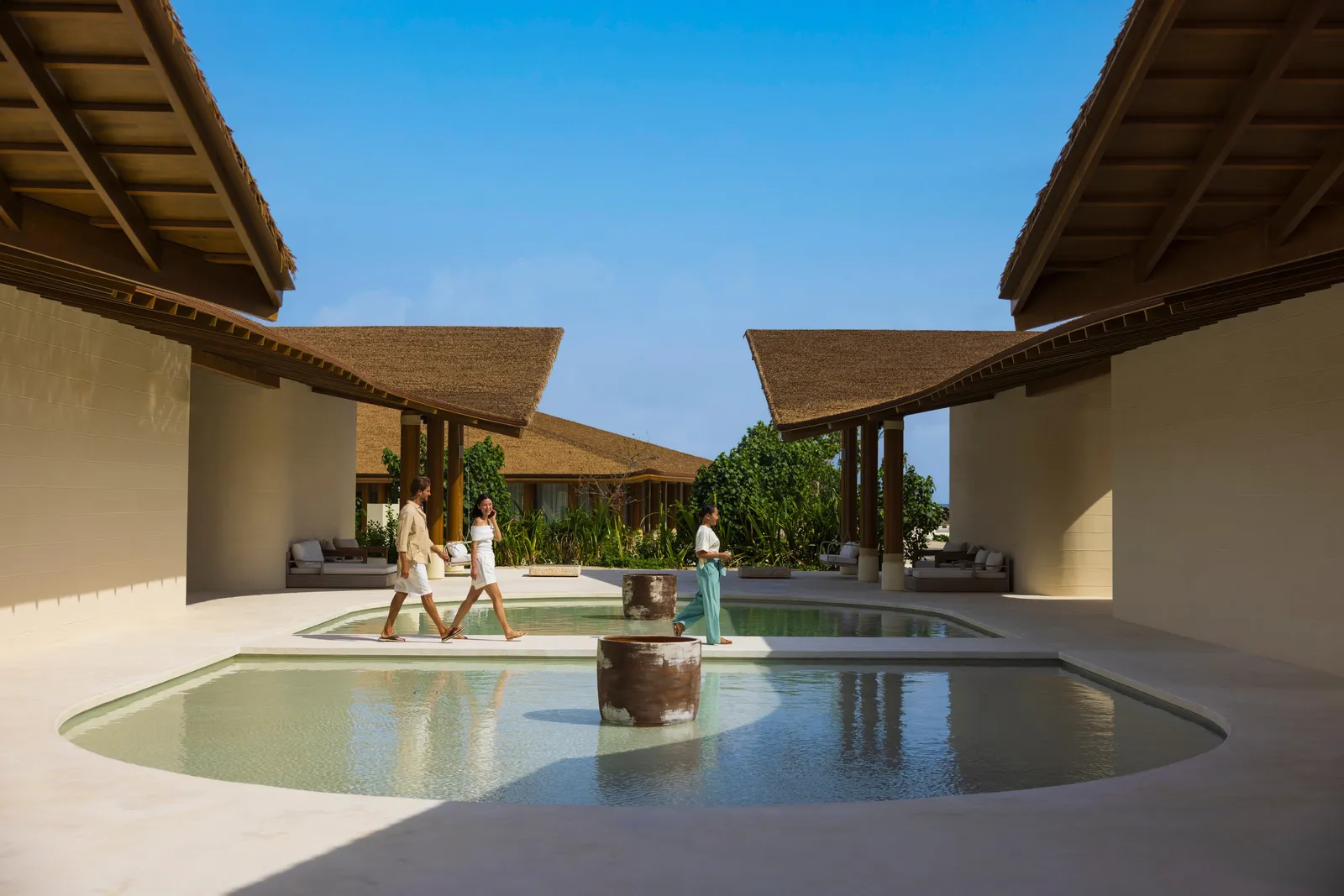 The SPA Cenvaree Retreat courtyard: shallow pools under thatched roofs, 3 people walking through