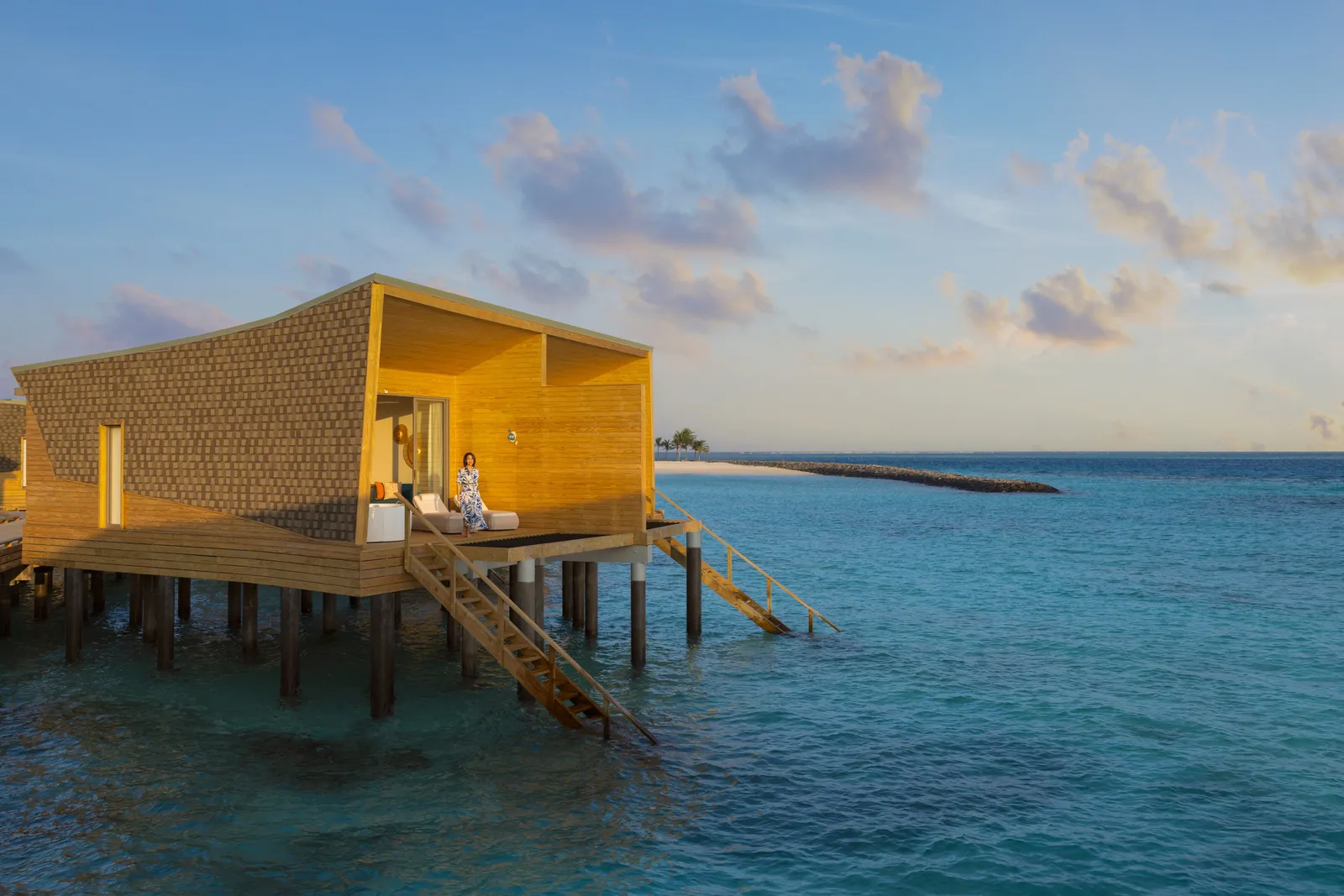 A pale-timber water villa at dusk, its ladder dropping into the lagoon