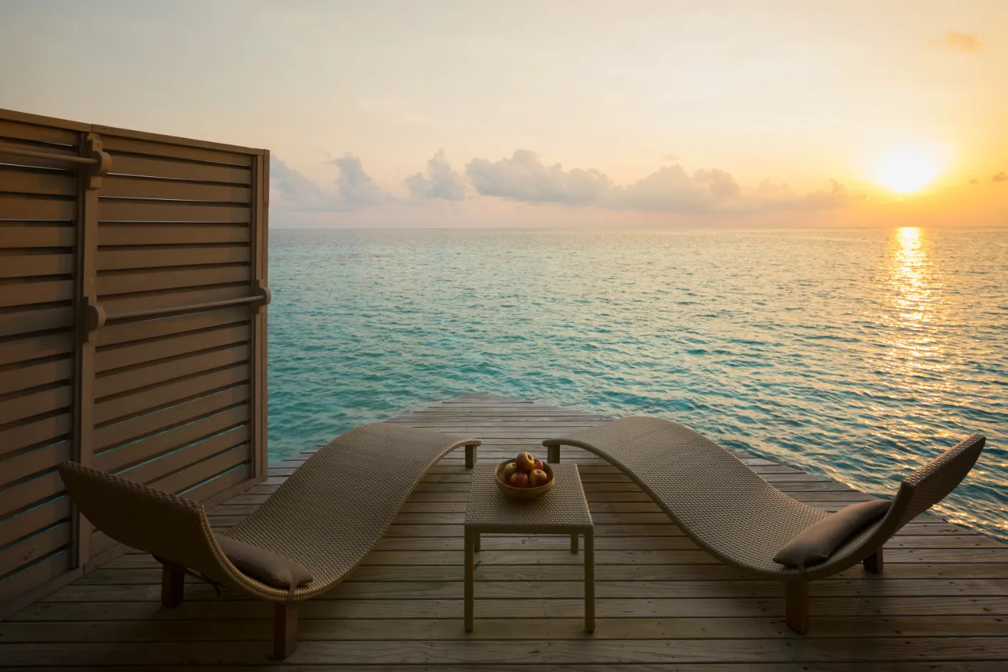 The deck of a Sunset Overwater Villa at sunset, 2 loungers facing the sea.