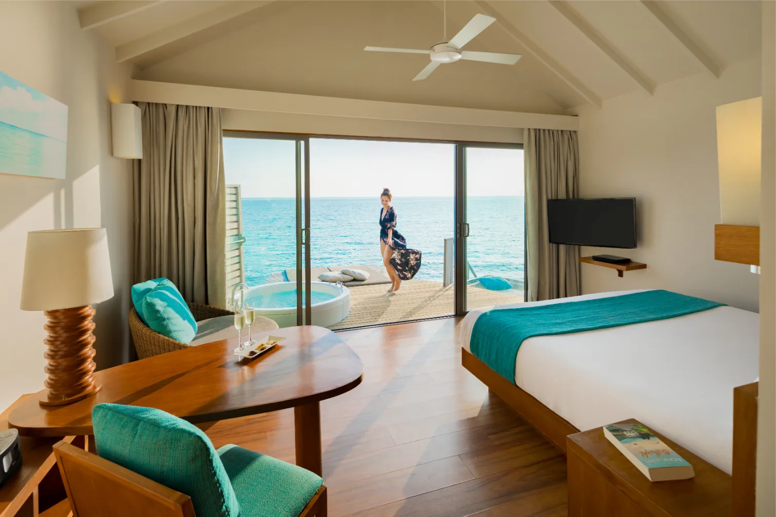 A Sunset Overwater Villa with Swirl Pool: the bed, the table and, beyond the glass, the deck tub where a guest looks out to sea.