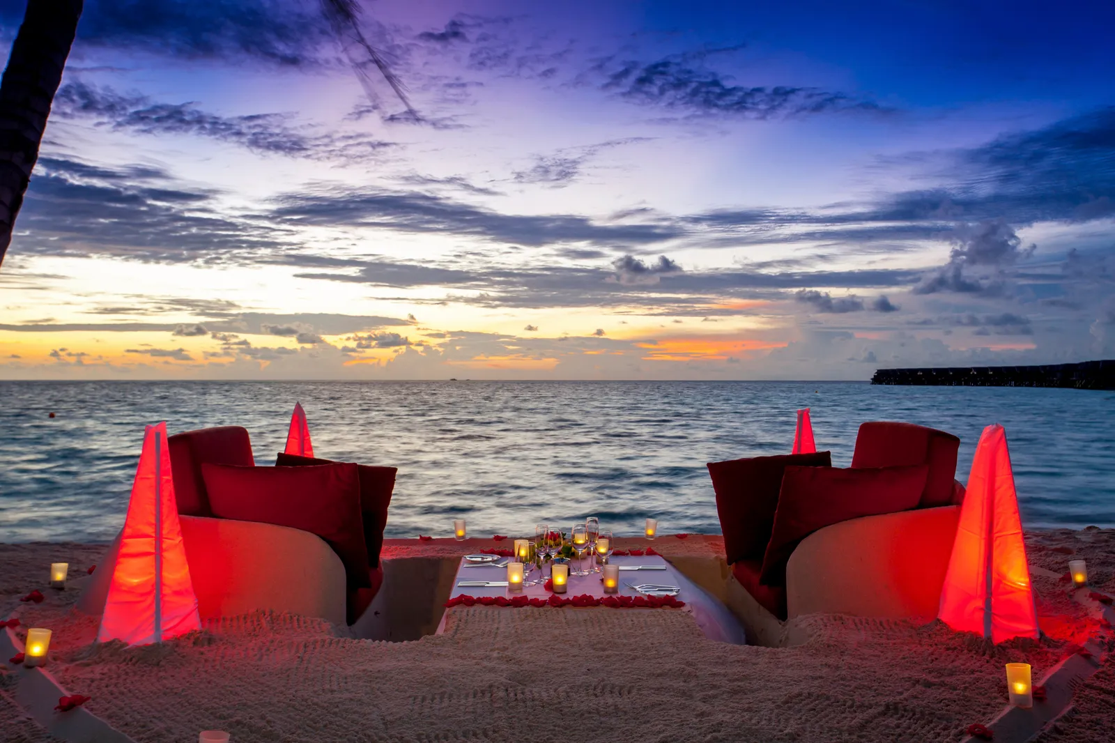A sand-sofa dinner on the shore, with red lanterns and a sunset sky.