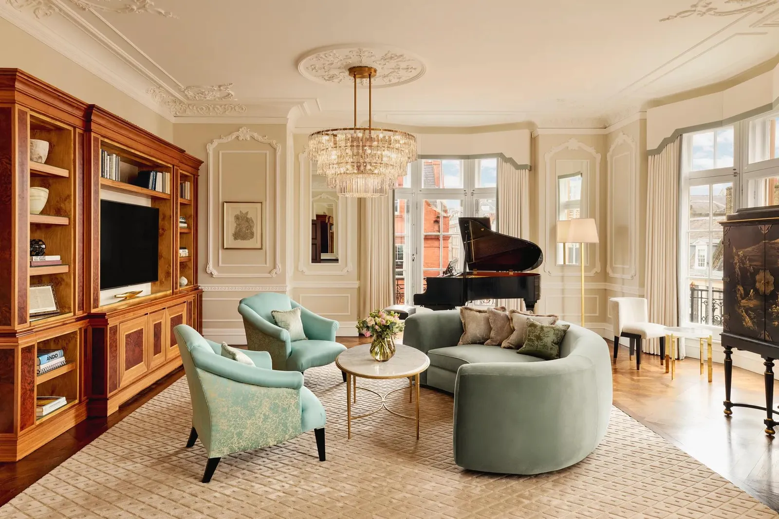 Elegant living room with a curved mint-green sofa, 2 teal armchairs, and a grand piano by tall windows. The room features ornate wall panelling, a crystal chandelier, and built-in wooden cabinetry with books and a television.