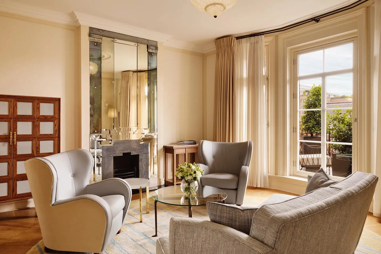 Elegant sitting area with grey armchairs and a matching sofa around a glass coffee table topped with flowers, set beside large windows and a mirrored fireplace reflecting warm natural light.