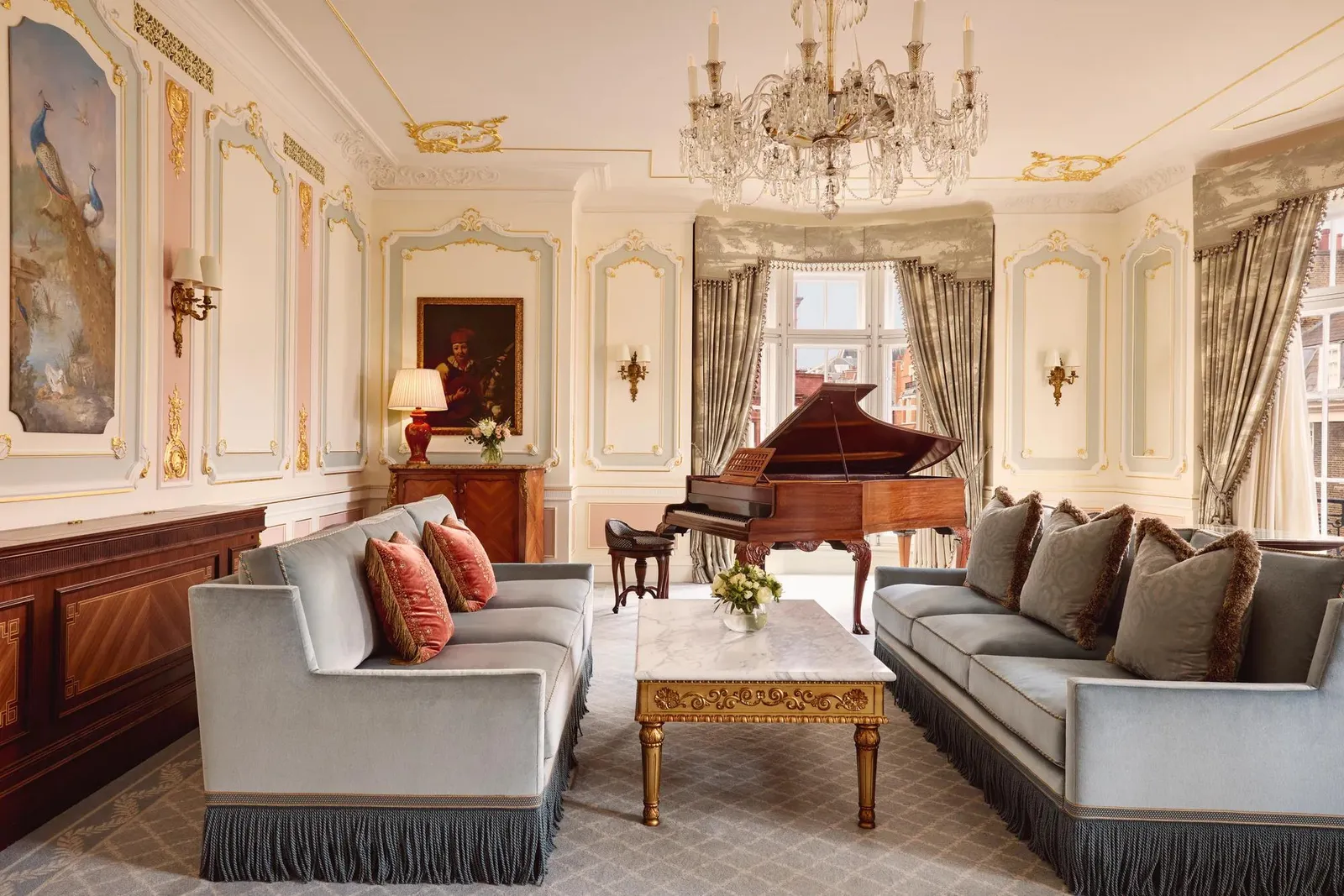 Opulent living room with 2 blue fringed sofas facing each other, a gold-detailed marble coffee table, and a grand piano beside tall curtained windows, illuminated by an ornate crystal chandelier.