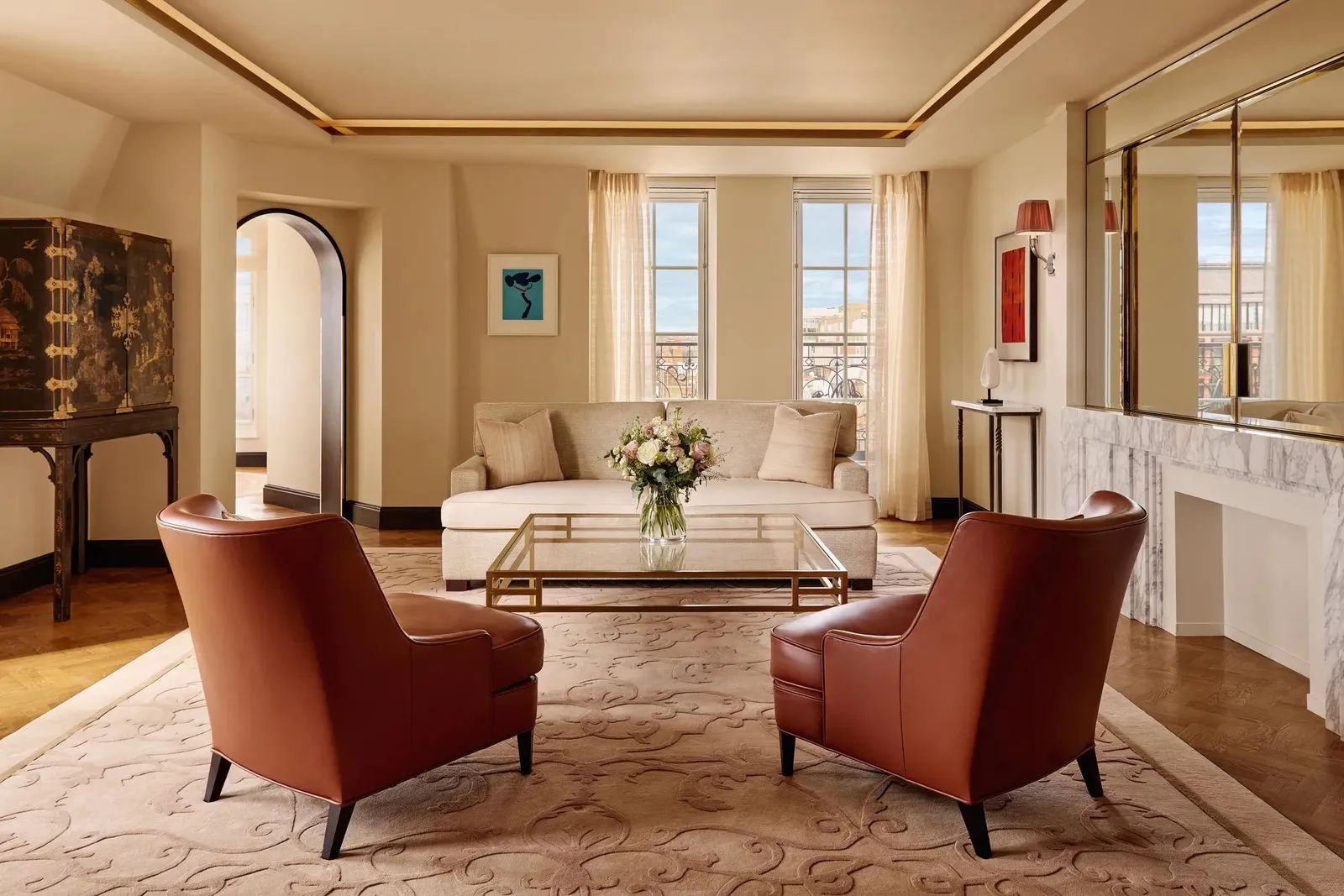 Bright living room with 2 brown leather armchairs facing a cream sofa, a glass coffee table with flowers, and large windows framed by sheer curtains, decorated in warm neutral tones with modern artwork.