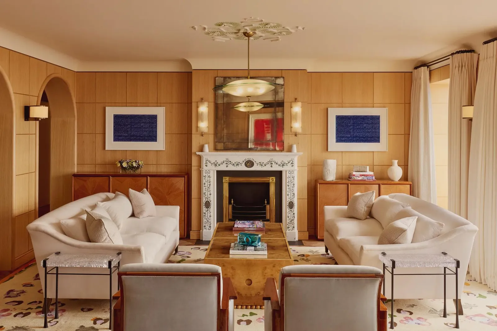 Elegant living room with 2 cream sofas facing each other, a marble fireplace with decorative tiles, and wood-paneled walls featuring framed abstract art in blue.