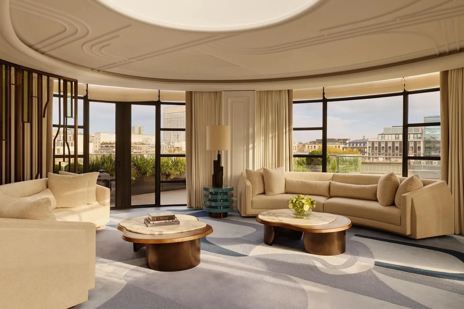 Spacious curved living room with large windows offering city views, featuring cream sofas, marble-topped coffee tables, a patterned blue and beige rug, and soft natural light filtering through full-length curtains.