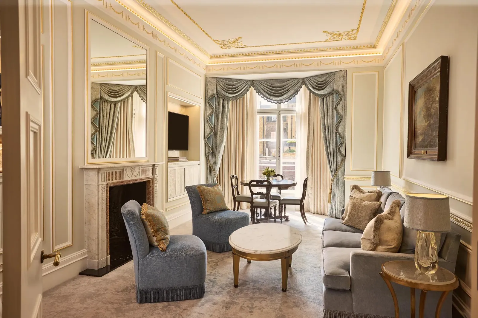 alt text Living room with blue upholstered seating, marble fireplace and ornate draped windows overlooking the city, with a dining table set by the window.