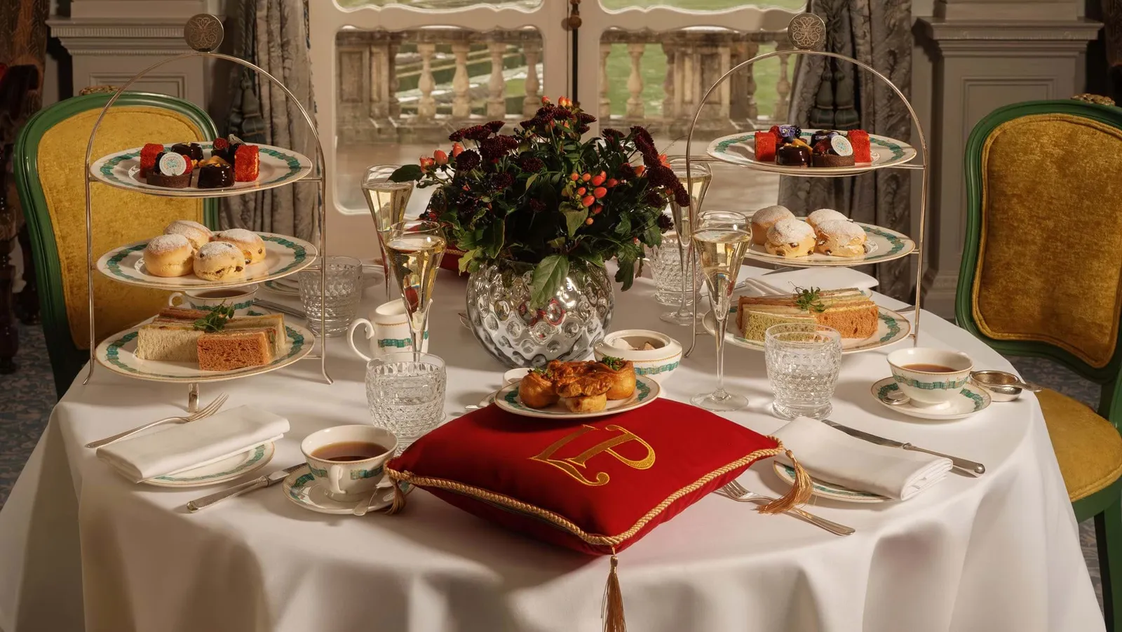 A tea table with cake stands, flutes and an embroidered red velvet cushion