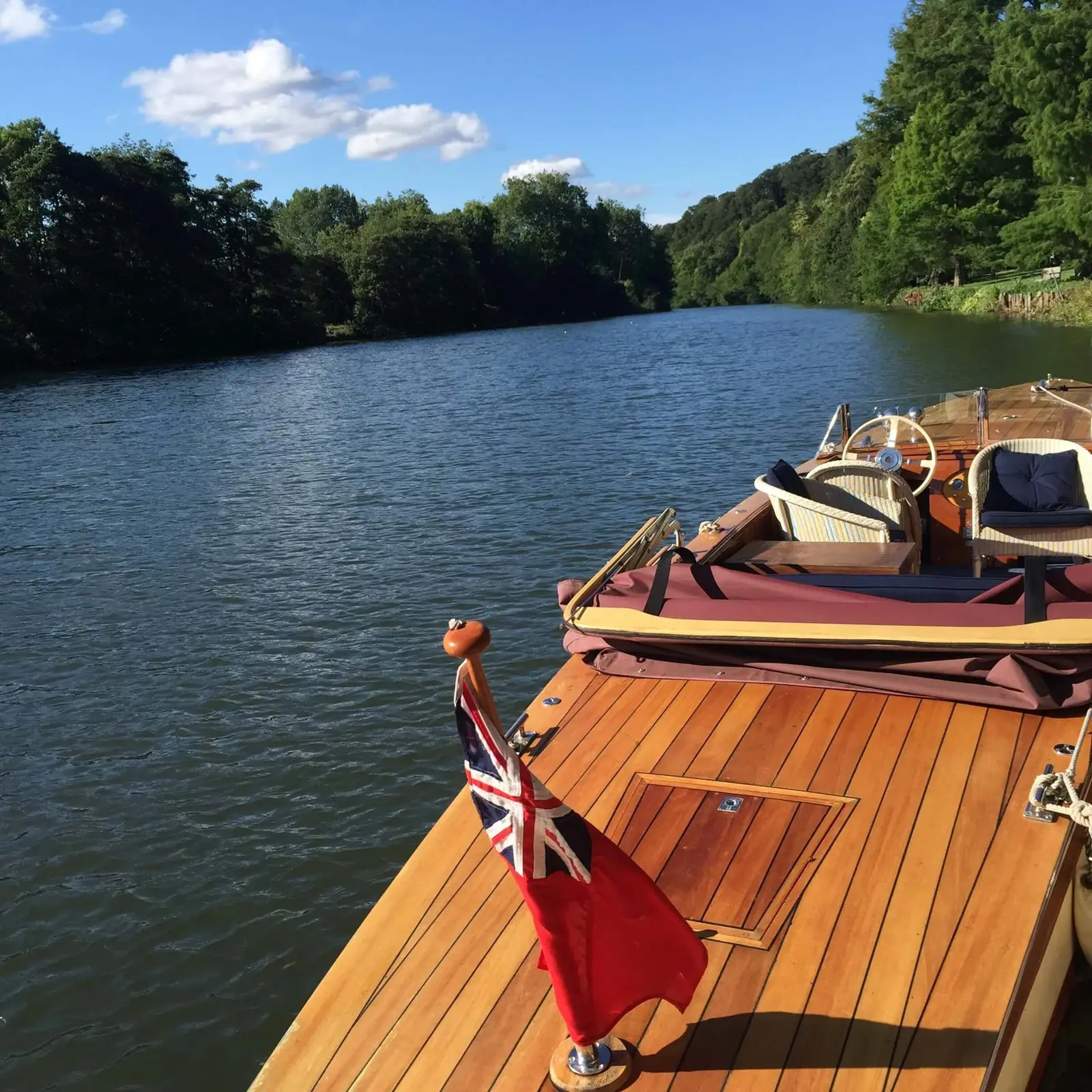 The wooden bow of a vintage launch flying the red ensign, on the Thames between the trees