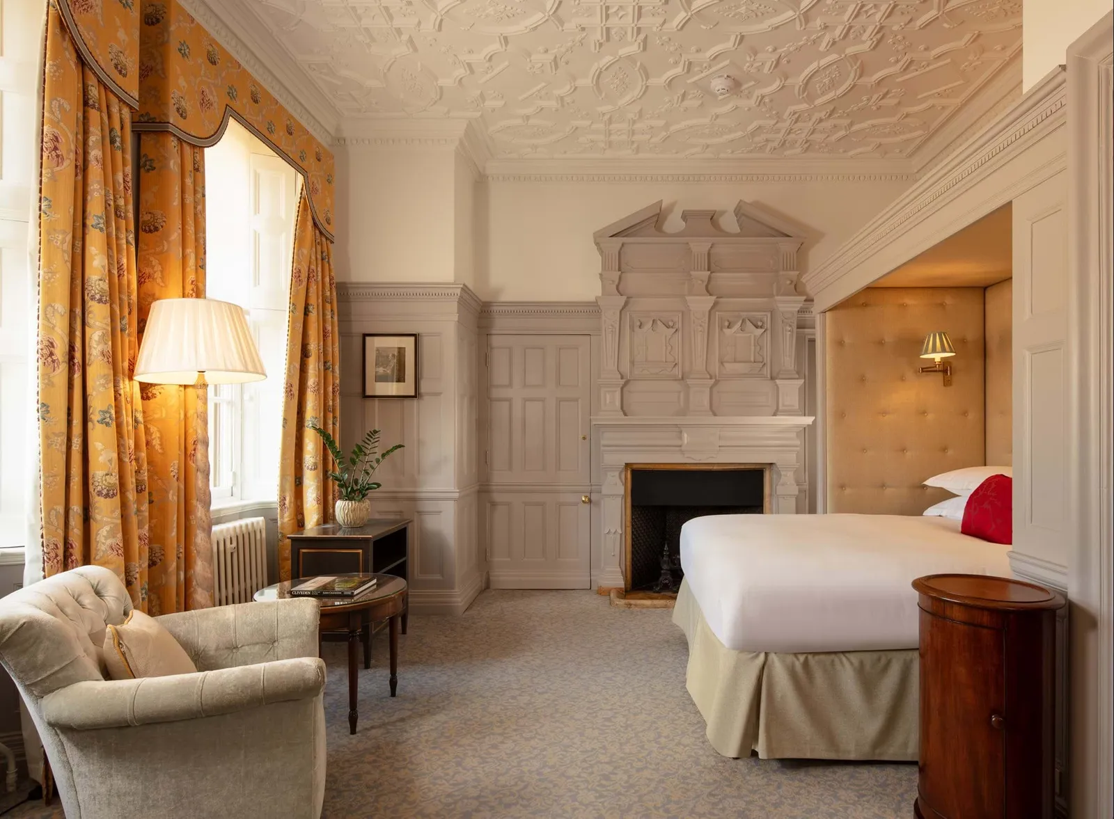 A Club Room: a bed with an upholstered headboard, a carved stone fireplace and floral curtains