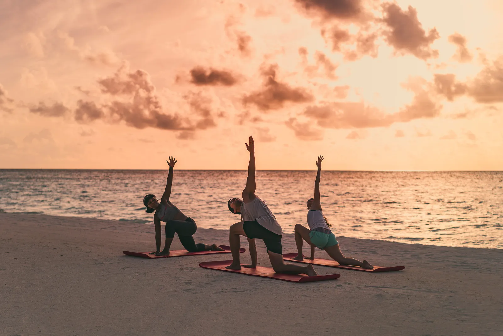 3 people doing yoga on the beach, on red mats, under the pink evening sky.