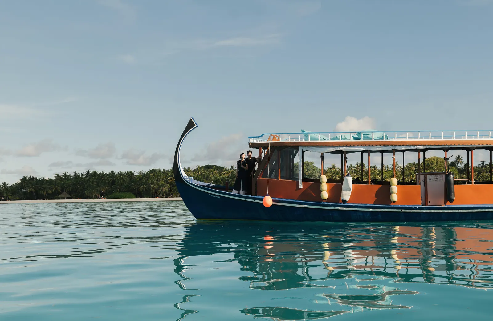 A dhoni, the traditional Maldivian boat with a tall prow, at rest on calm water with 2 people aboard.