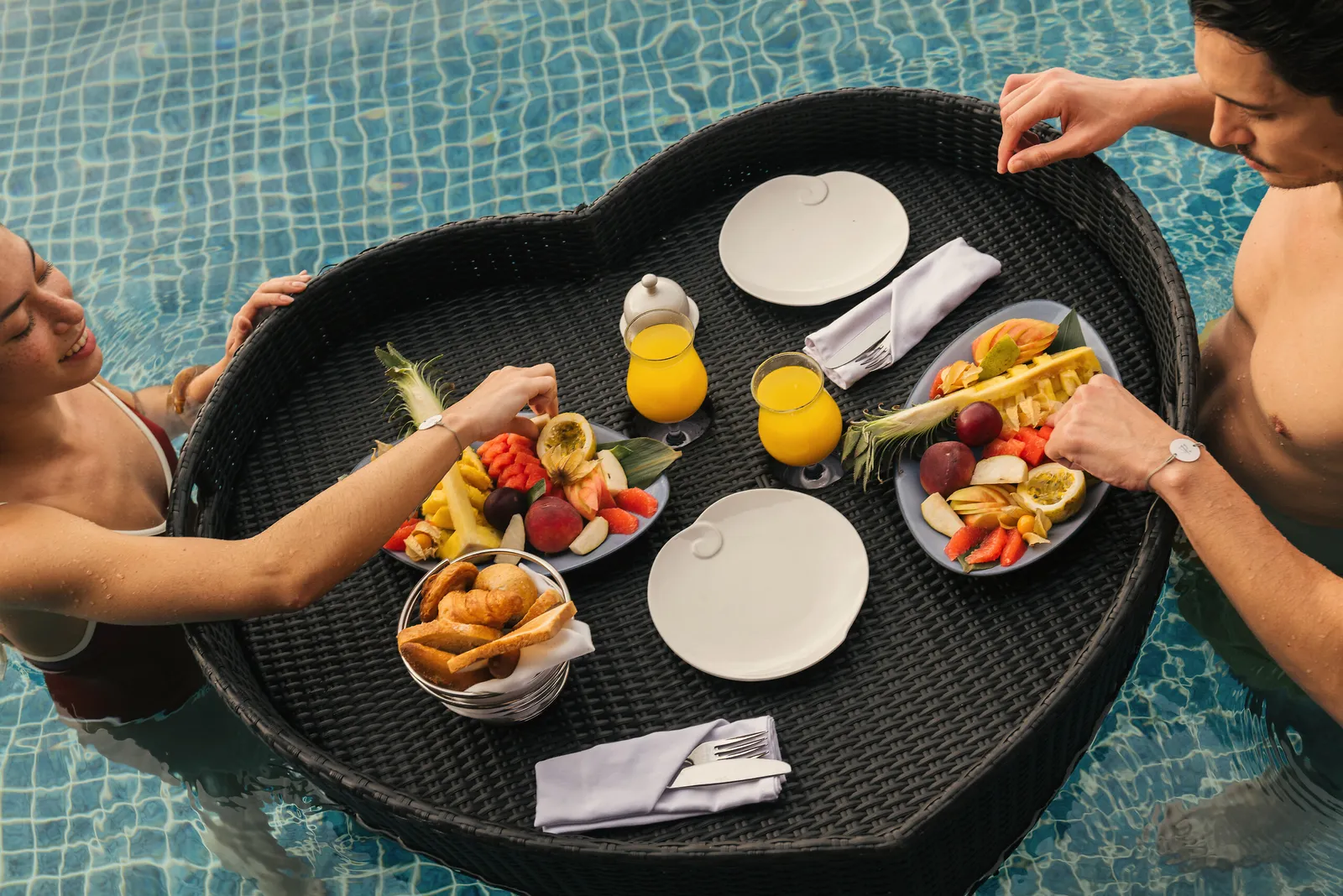 A couple in their villa pool with a heart-shaped floating tray of fruit, juice and pastries.
