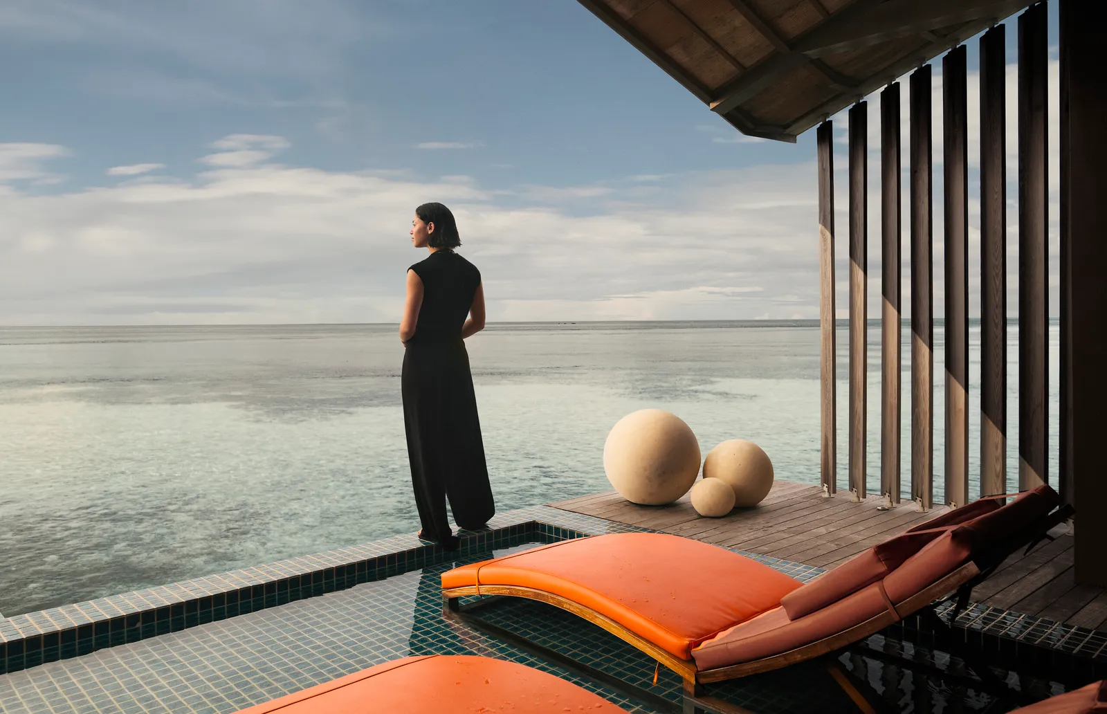 A woman standing at the edge of a water villa’s infinity pool, facing the calm sea, beside the orange loungers.