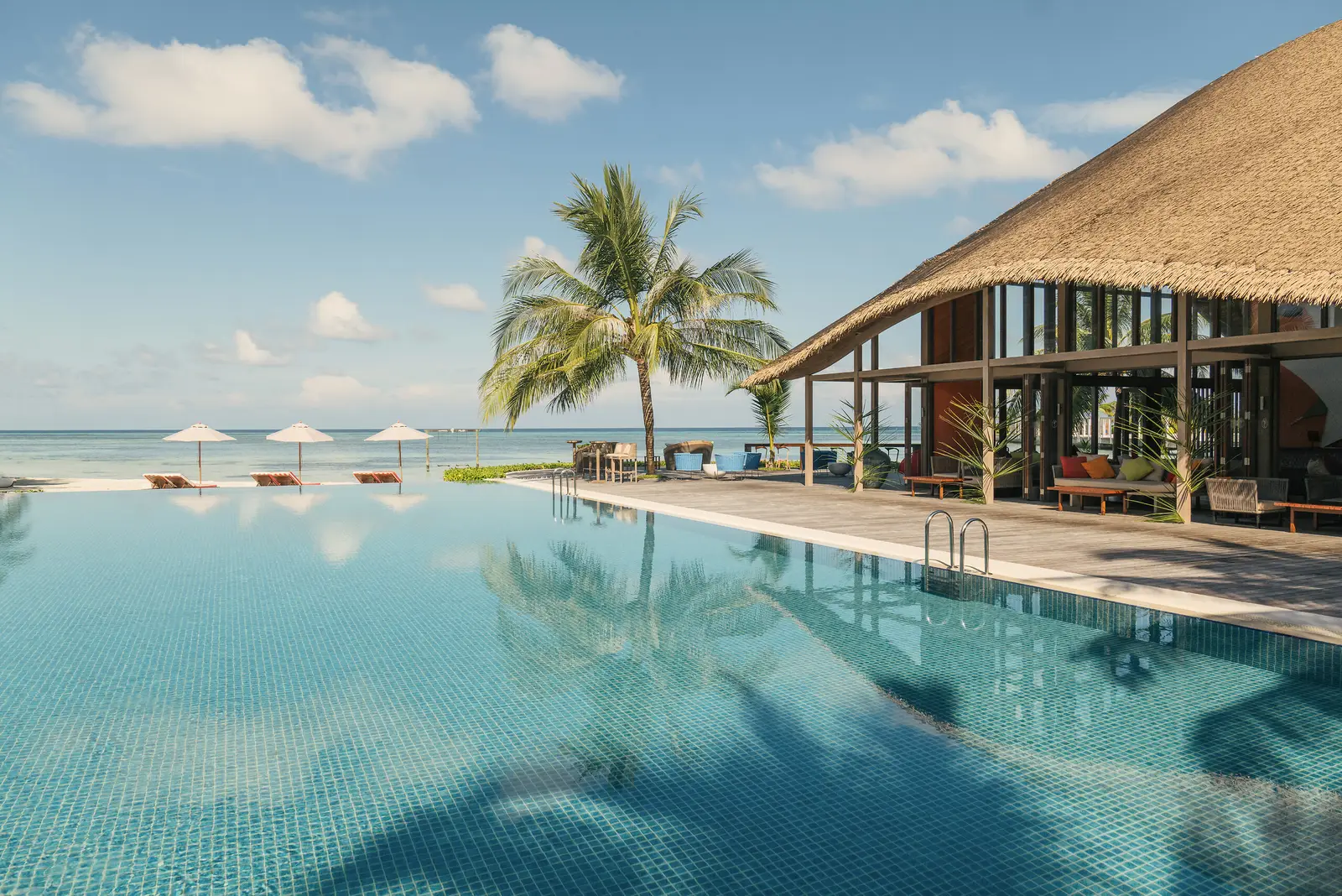 The main infinity pool, a palm reflected in the water, the thatched pavilion and the parasols on the beach.