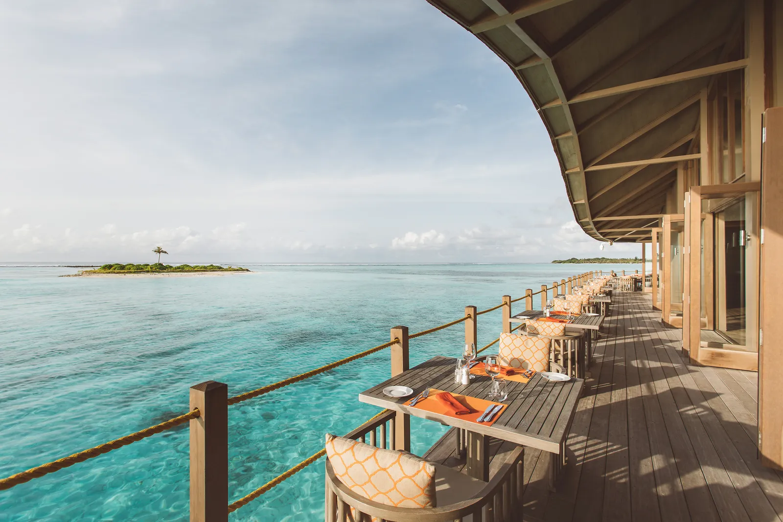 The Motu’s deck over the lagoon: tables laid along the rope rail and an islet with a single palm on the horizon.