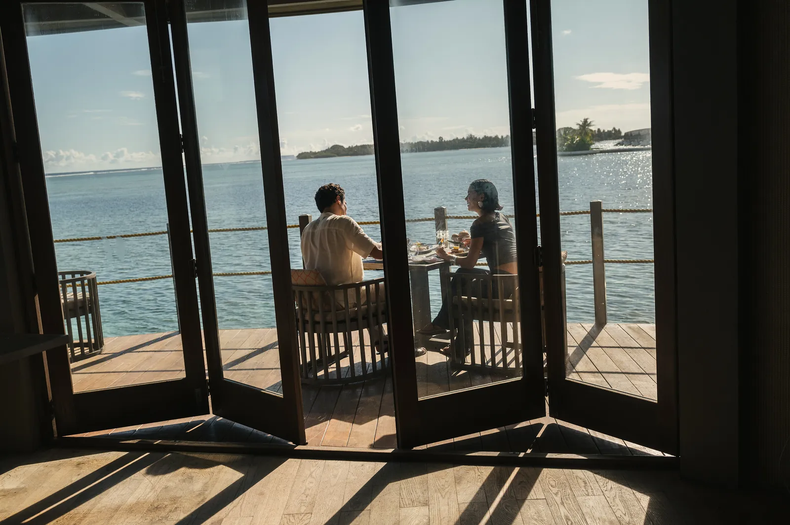 A couple at a table on the Motu’s deck, seen through the folding glass doors open onto the lagoon.