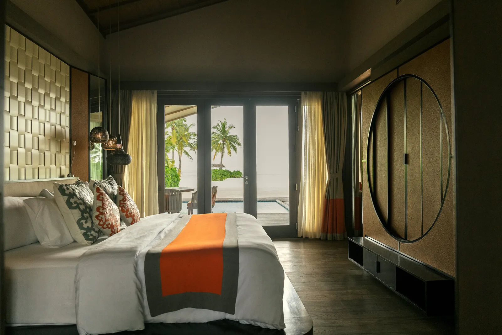 A sunset-side Beach Villa bedroom: the bed with its orange runner, the timber walls and the glass doors onto the garden.