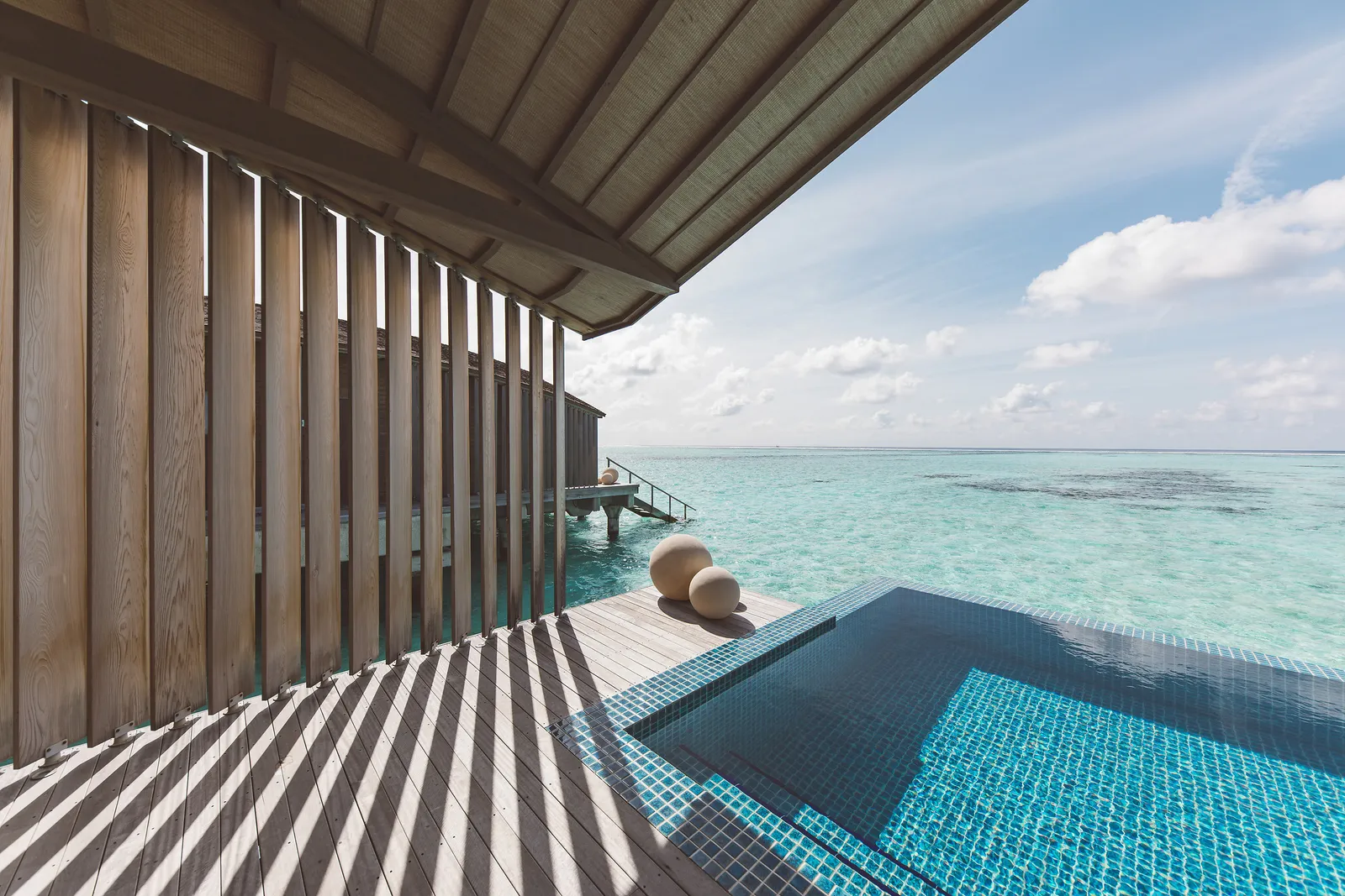A sunrise-side water villa’s deck: the infinity pool, the shaded timber slats and the open sea.