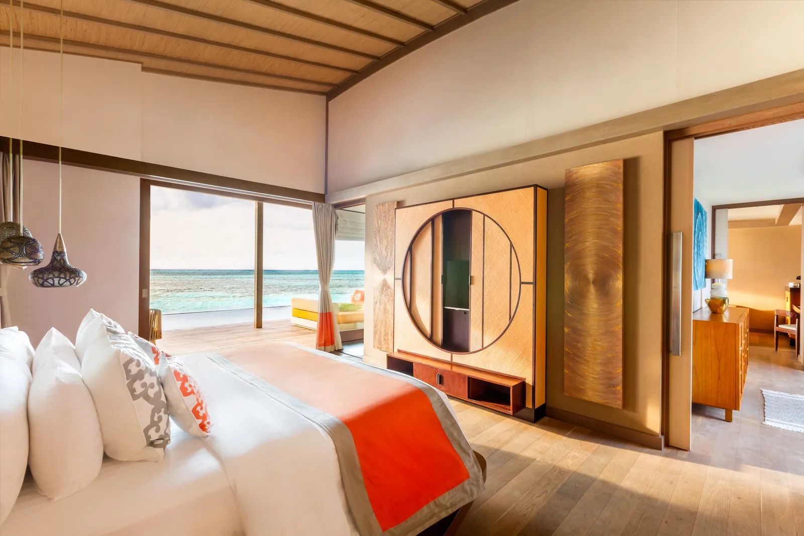 A sunset-side water villa bedroom, the bed facing the glass wall open onto the deck and the sea.